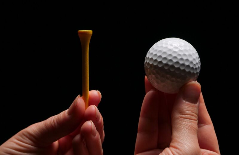 Two hands holding a wooden tee and a golf ball against a dark background