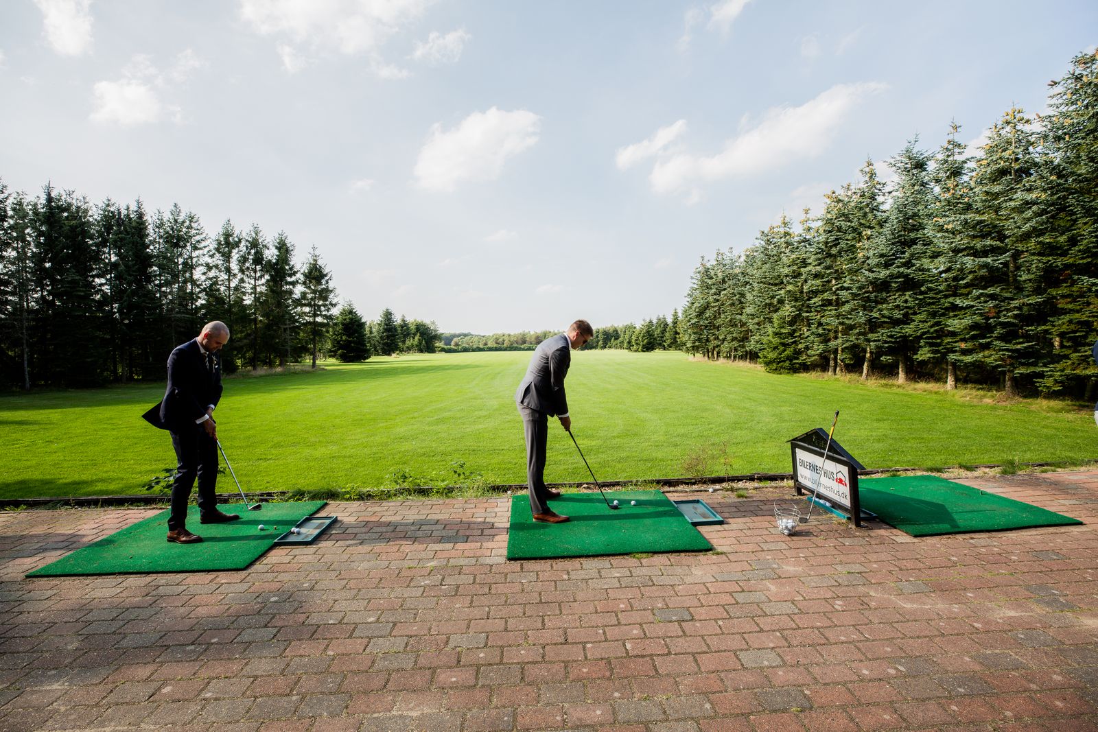 Two golfers hitting balls from mats on an outdoor driving range lined with trees