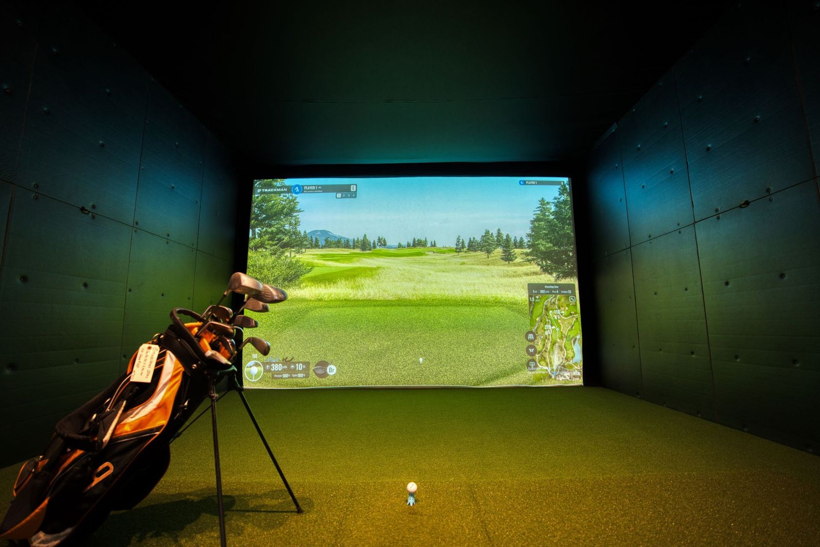 An indoor golf studio with a projection screen, a ball on a tee and a bag of clubs on a stand