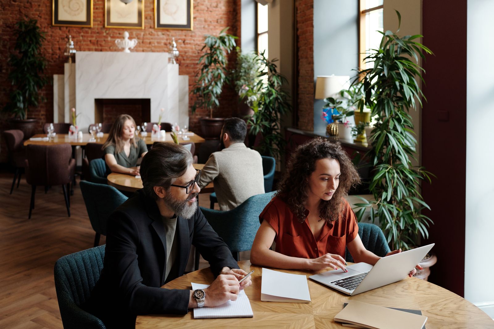 Two colleagues working on laptops and notes at a café table, with others talking behind them