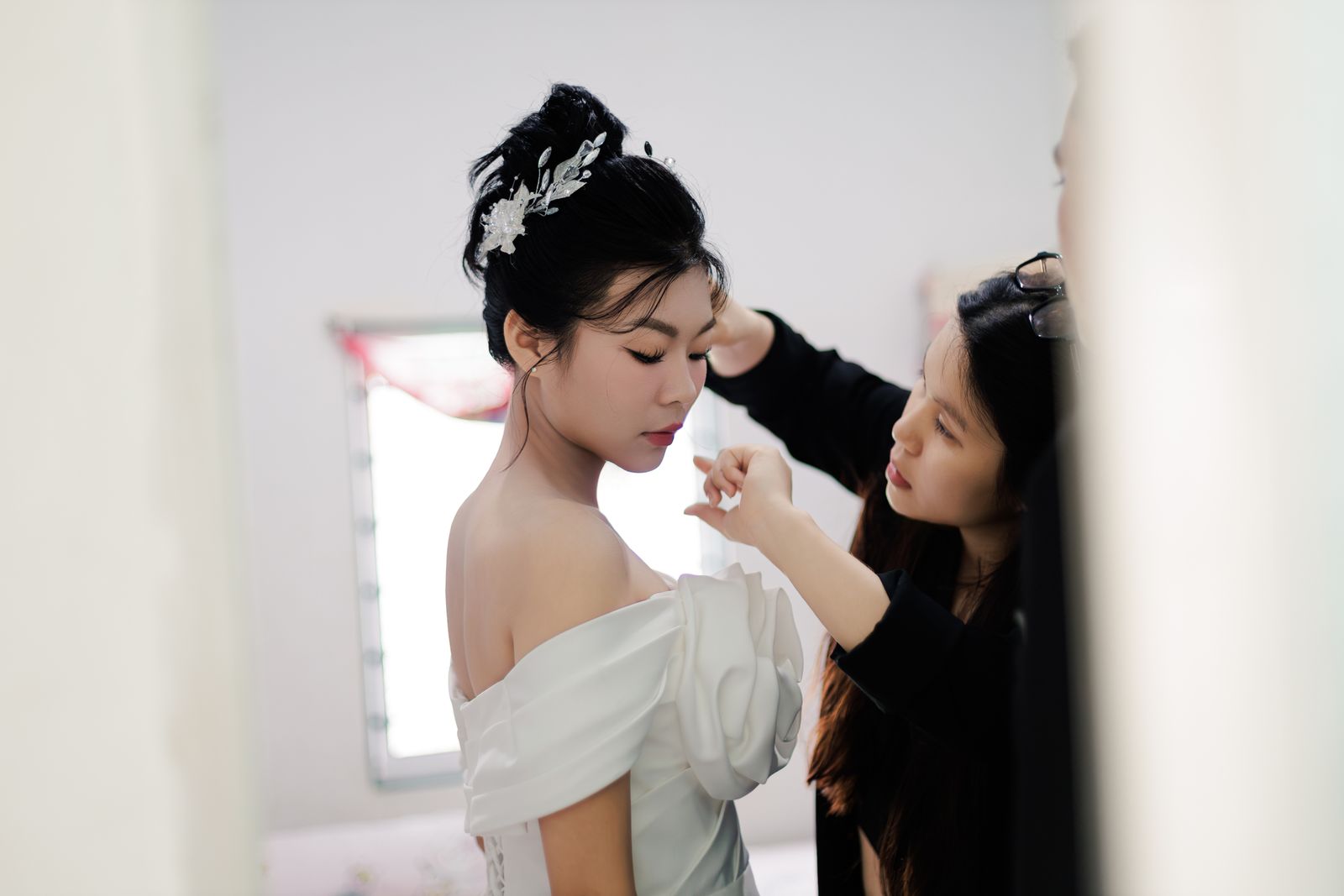 A makeup artist finishes a bride's makeup by a bright window