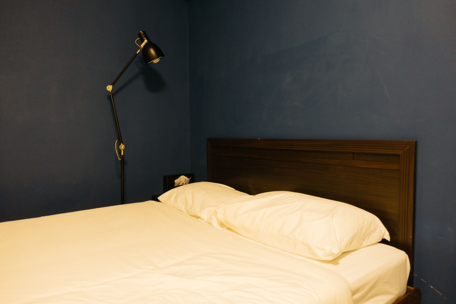 A made bed with cream linen against a dark blue wall, a black reading lamp above the headboard