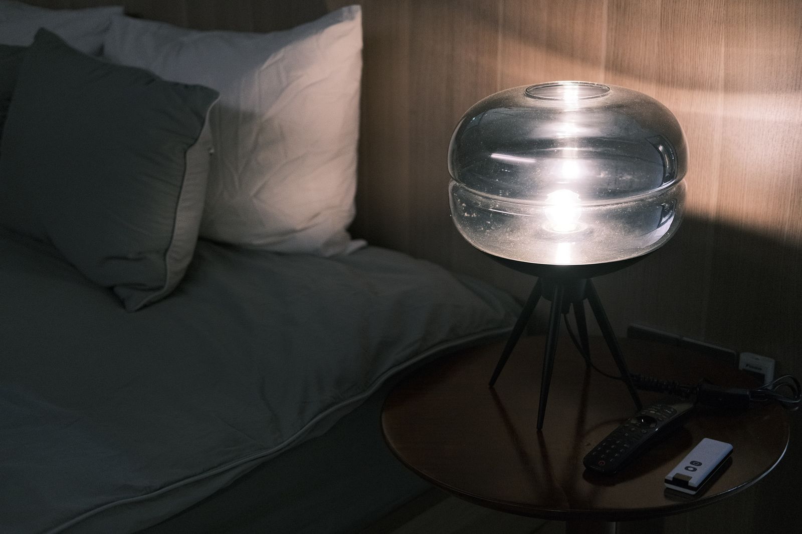 A glass bedside lamp glowing at night next to grey pillows