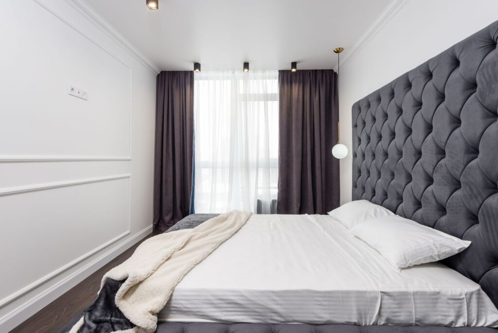 A bedroom with a tall tufted headboard, a made bed and long dark curtains