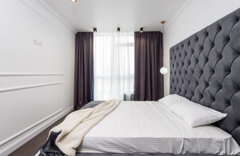 A bedroom with a tall tufted headboard, a made bed and long dark curtains