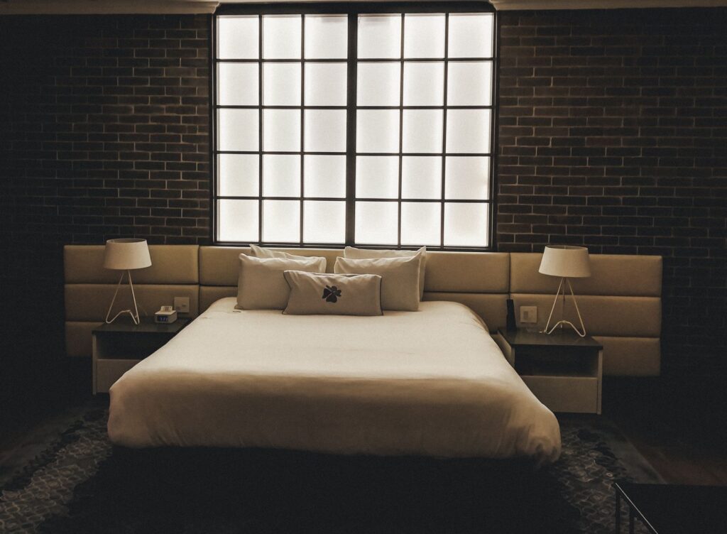 A bed with white linen between two lit bedside lamps in a dark room with a brick wall and a tall window