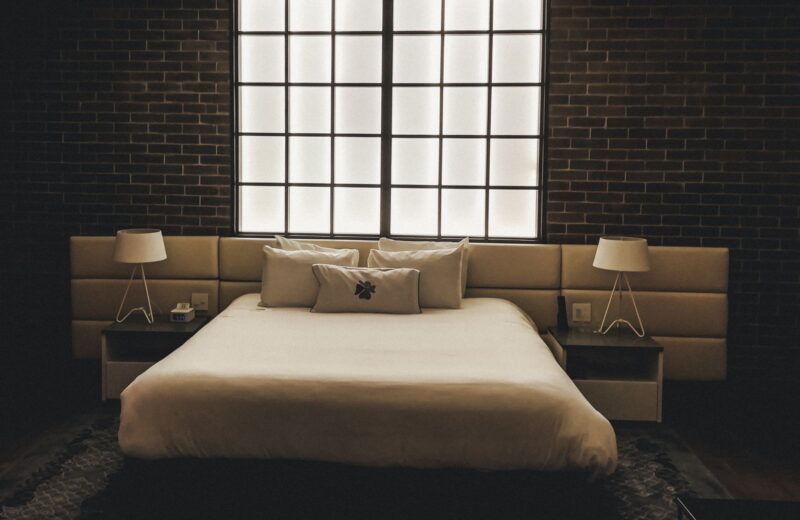 A bed with white linen between two lit bedside lamps in a dark room with a brick wall and a tall window