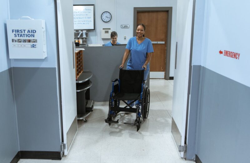 A nurse pushing an empty wheelchair along a hospital corridor