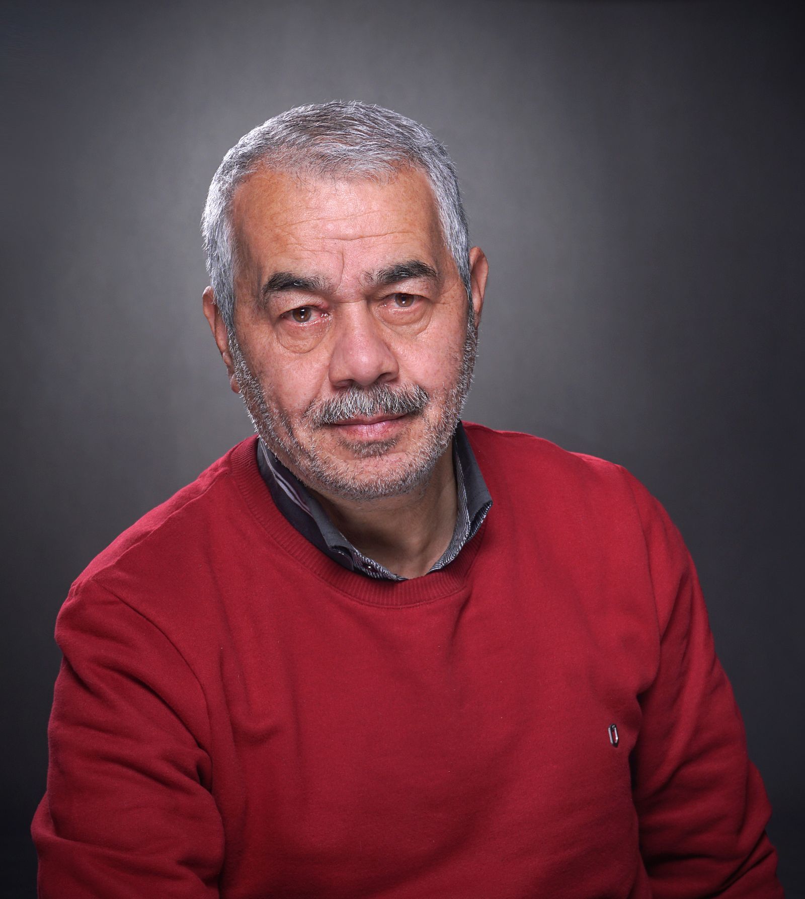 A man with white hair and a grey beard in a red sweater, photographed against a dark grey background