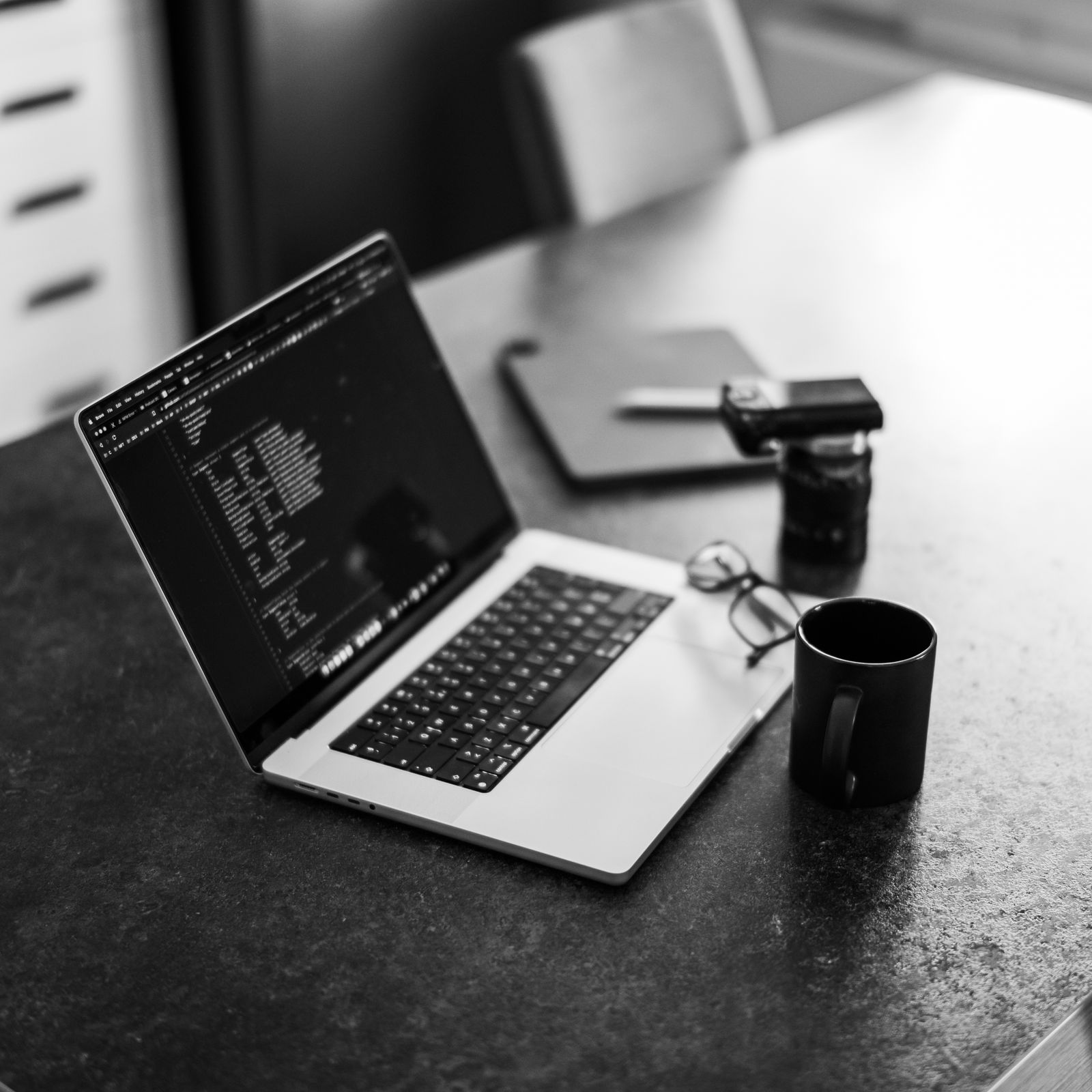 Black and white photo of a laptop showing code, beside a mug, glasses and a phone on a stone table