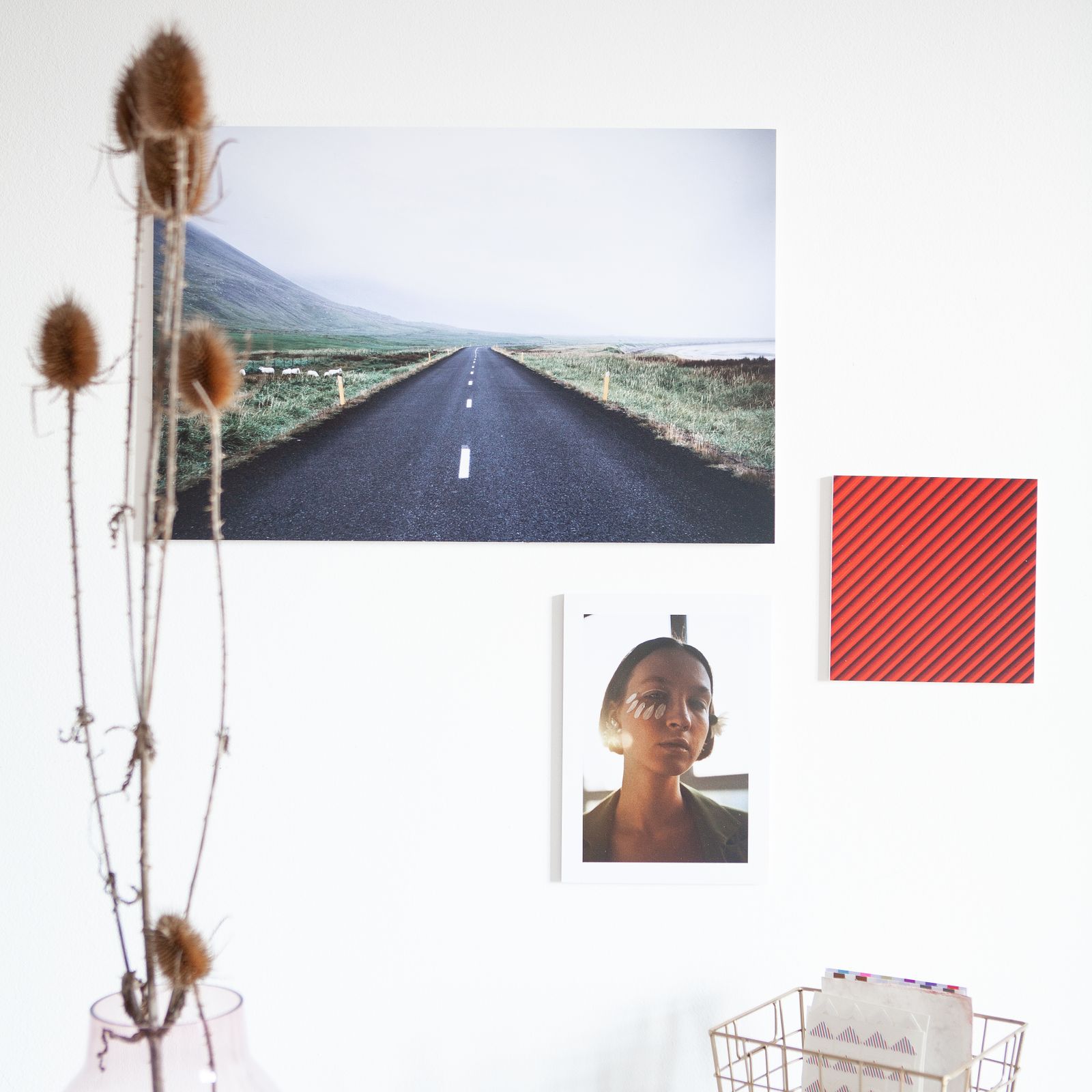 Prints pinned on a white wall: a road through green hills, a portrait and a small red square, with dried teasels in front
