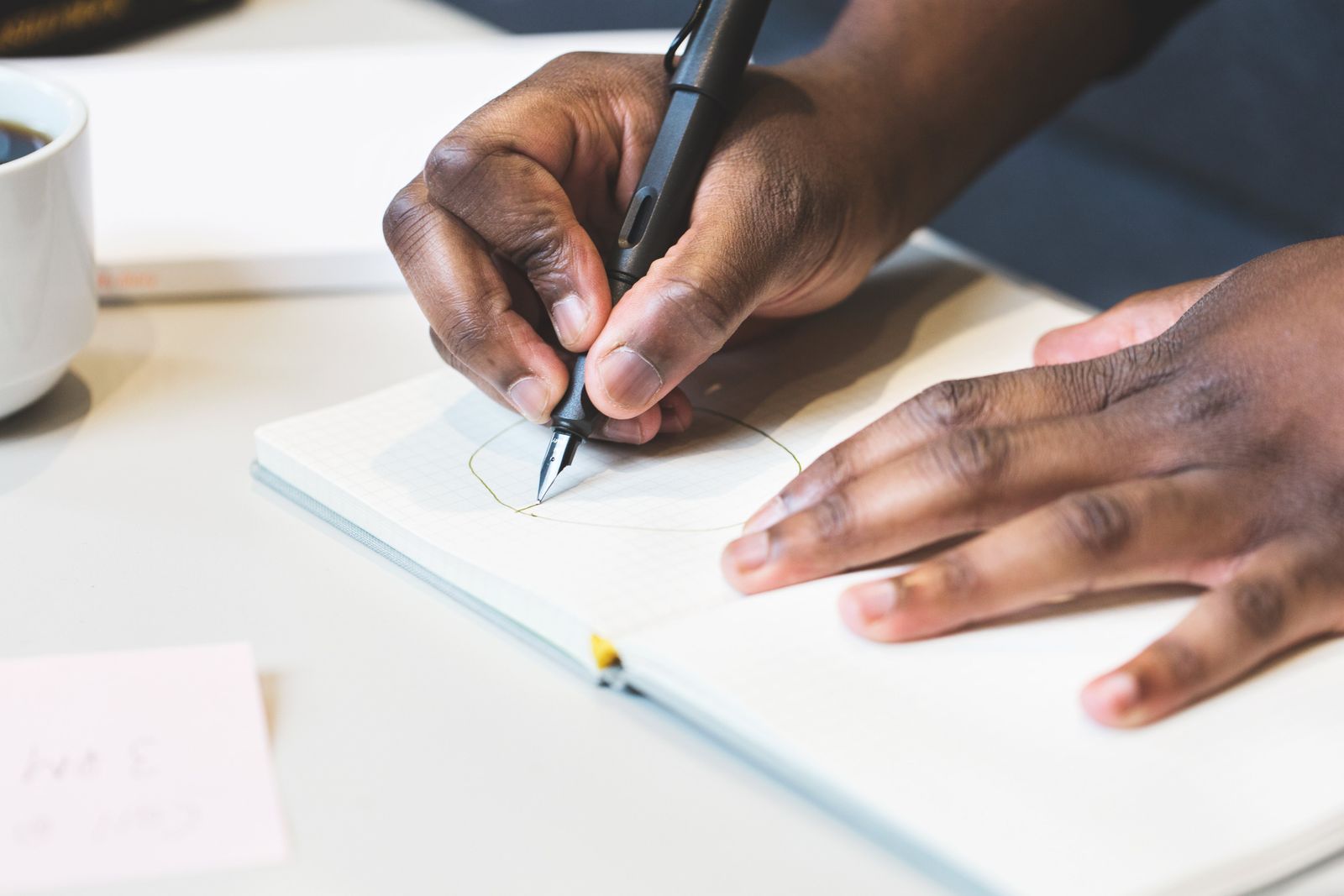 A hand writing in a notebook with a fountain pen, a cup of coffee beside it
