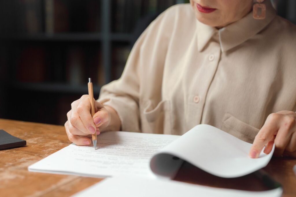 A woman at a wooden desk reading printed pages with a pencil in her hand