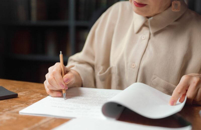 A woman at a wooden desk reading printed pages with a pencil in her hand