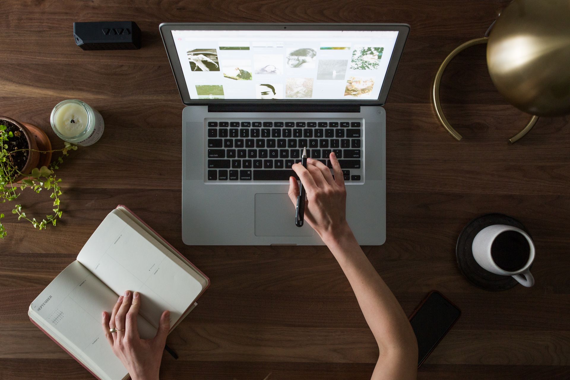 A dark wooden desk seen from above with a laptop, an open notebook and a cup of coffee