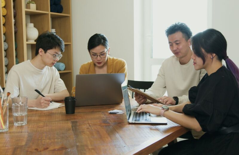 Four colleagues at a wooden table in a bright office, one reading from a notebook