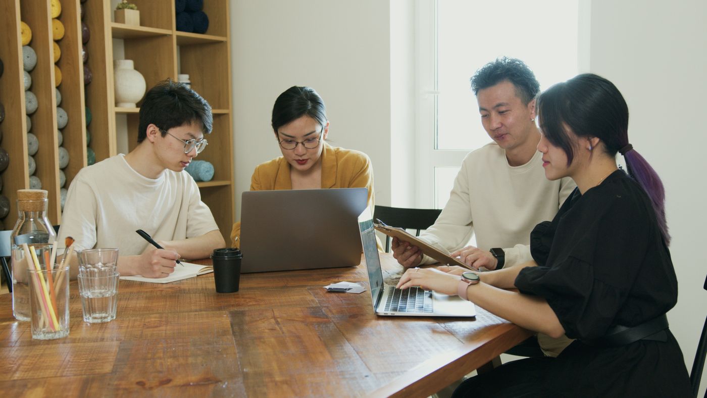 Four colleagues at a wooden table in a bright office, one reading from a notebook