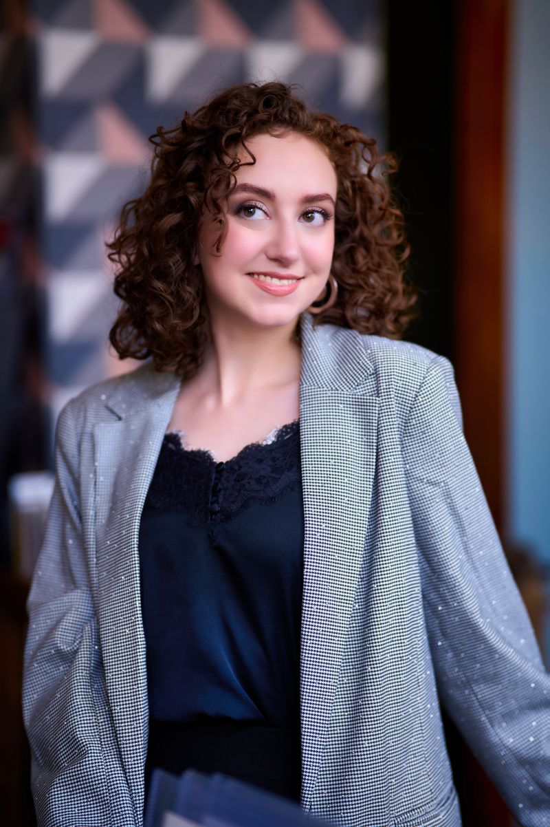 Portrait of a woman with curly hair in a grey blazer, smiling in an office