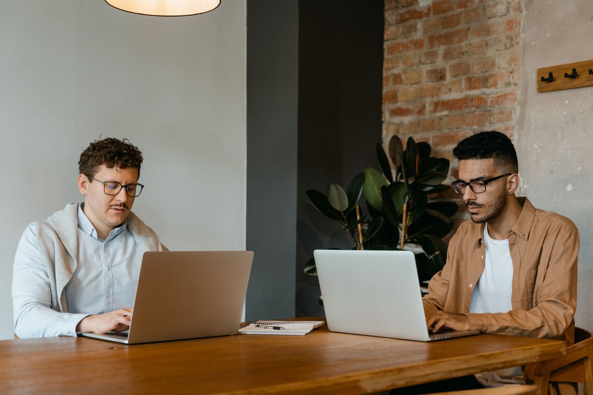 Two people working on laptops at a shared wooden desk by a brick wall
