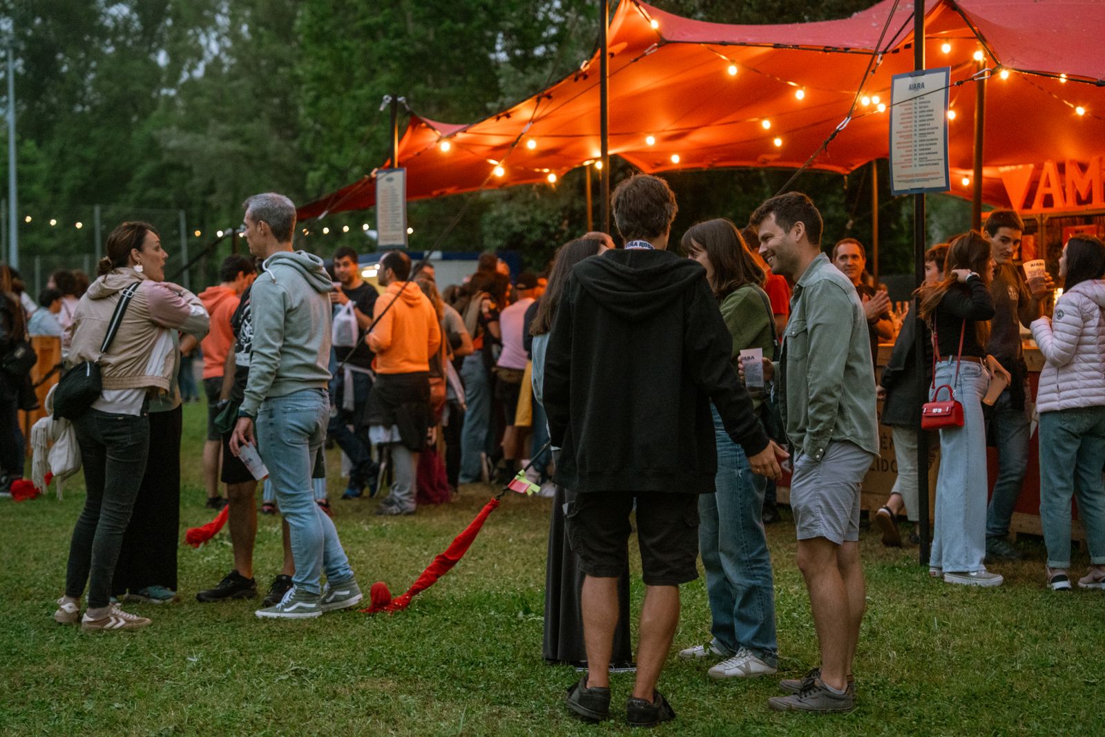 People standing and talking on the grass under a red festival tent