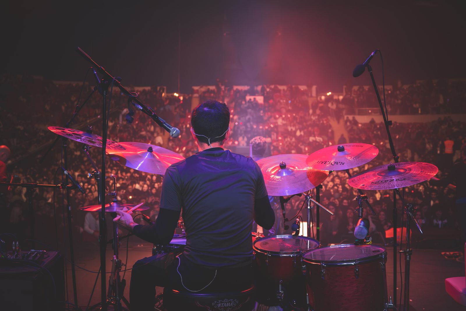 A drummer seen from behind, playing to a packed crowd under stage lights