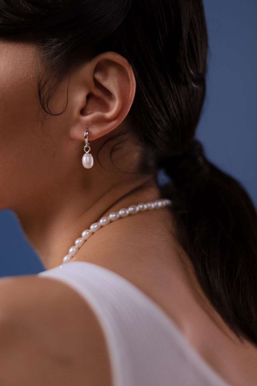 A pearl drop earring and a pearl necklace worn against a navy background, seen from behind