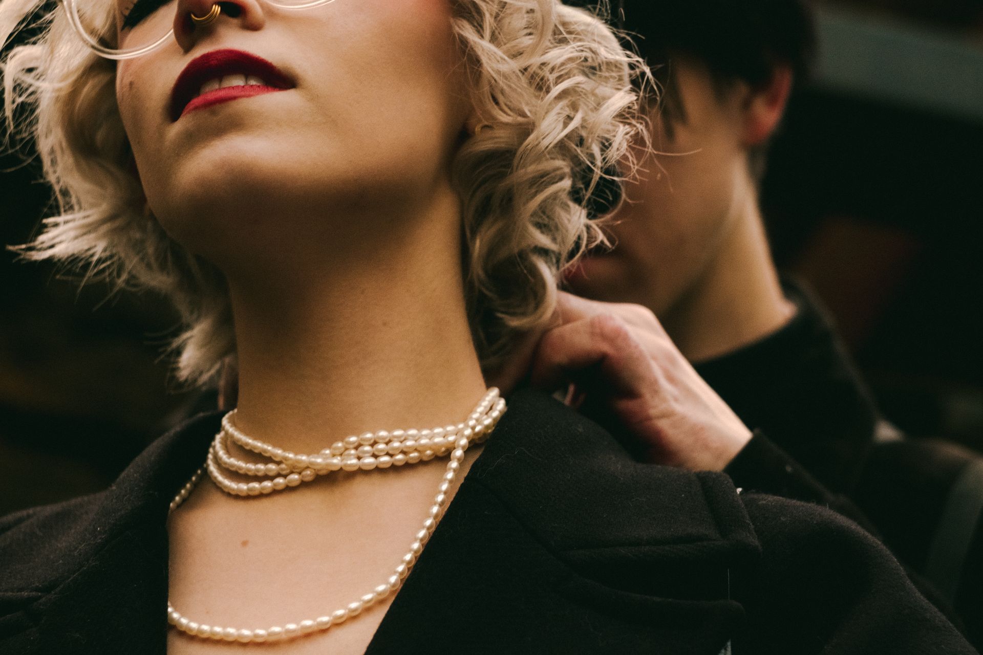 A woman in a black jacket having a long pearl necklace fastened at her neck, warm light