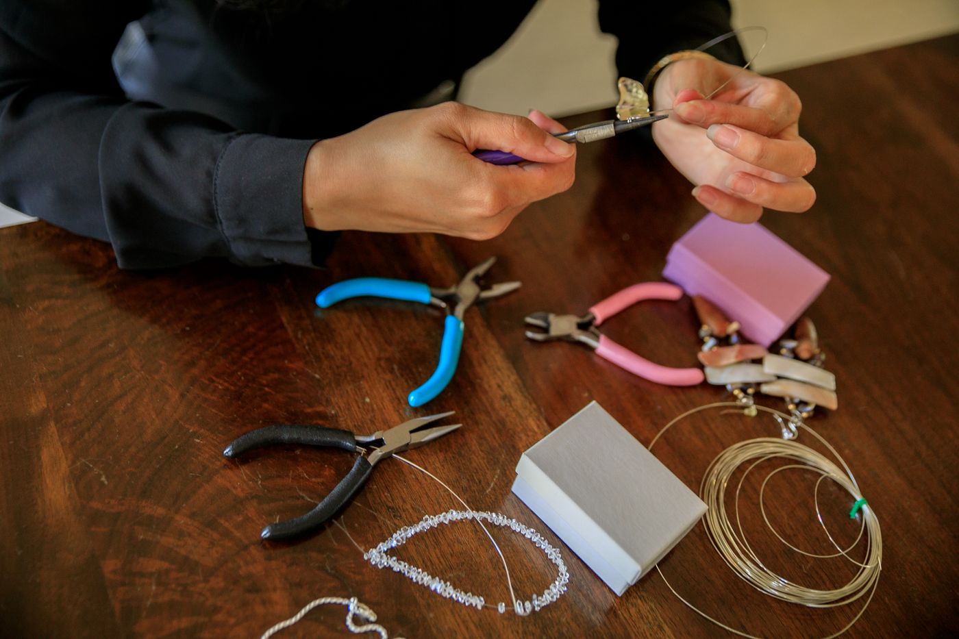 Hands with pliers threading a necklace on a dark wooden workbench, tools around