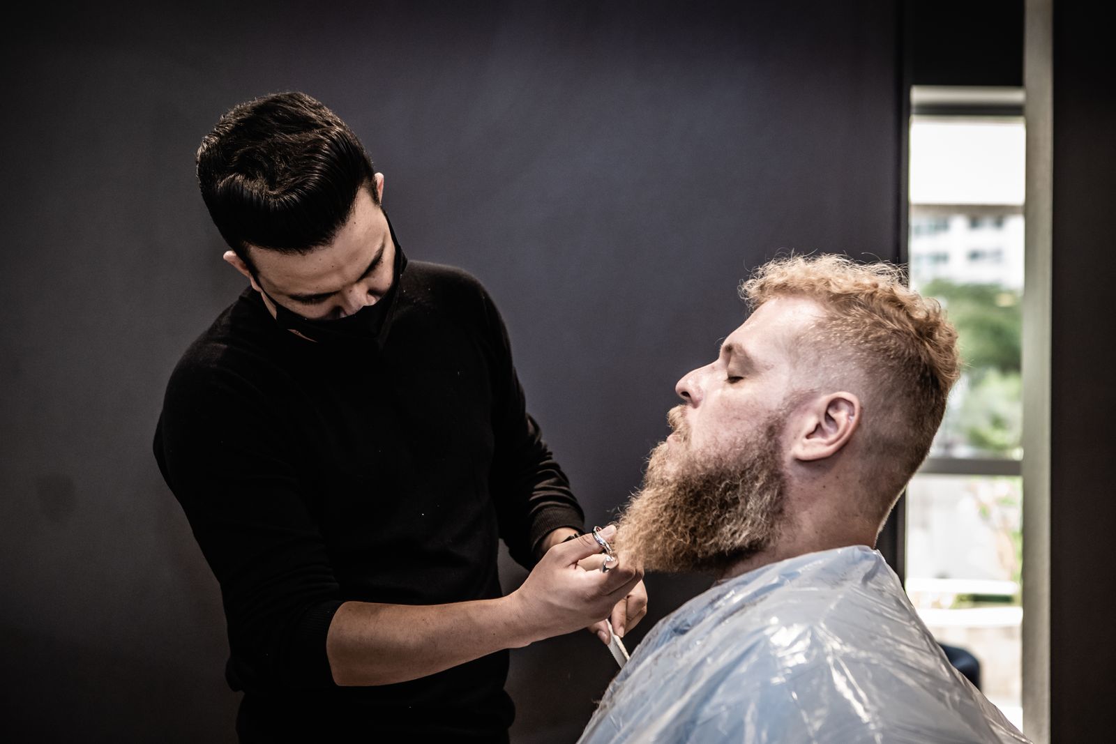 A barber trimming a client's beard in a dark, quiet shop