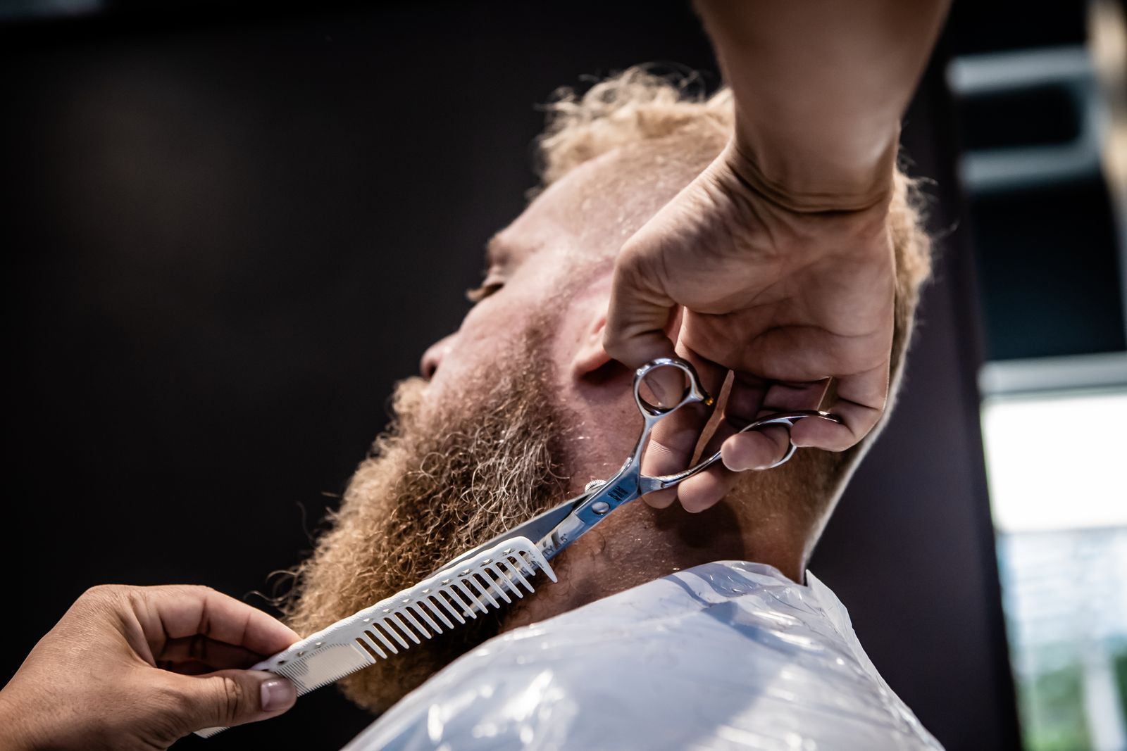 A barber shaping a long beard with scissors and a comb