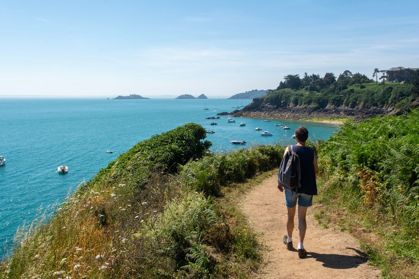 A woman seen from behind walking a coastal path above a blue bay
