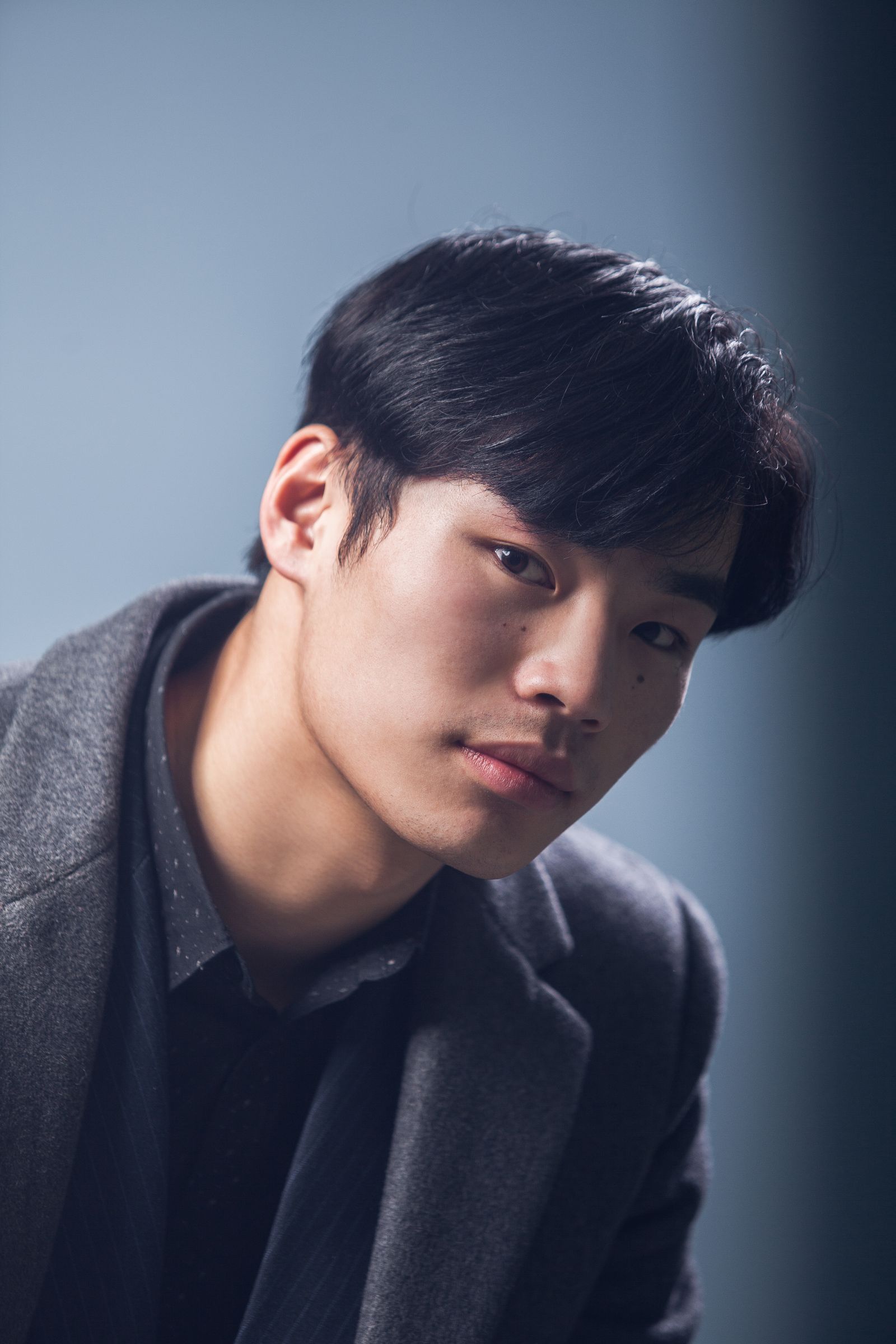 Studio portrait of a young man in a grey coat against a blue-grey backdrop, lit from the left