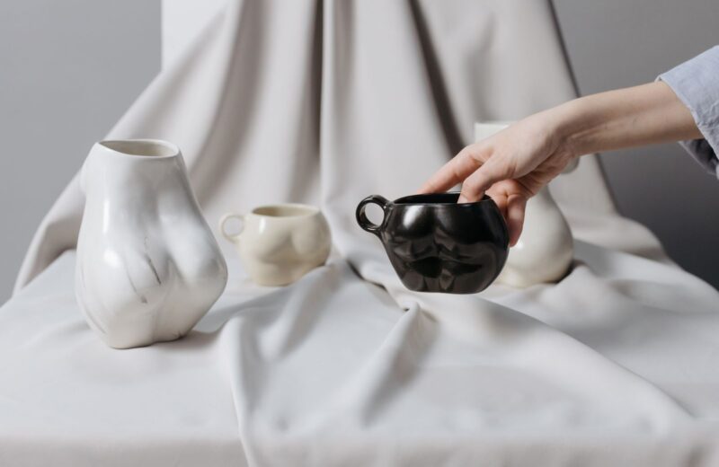 A hand placing a black ceramic teapot beside white vases on draped linen