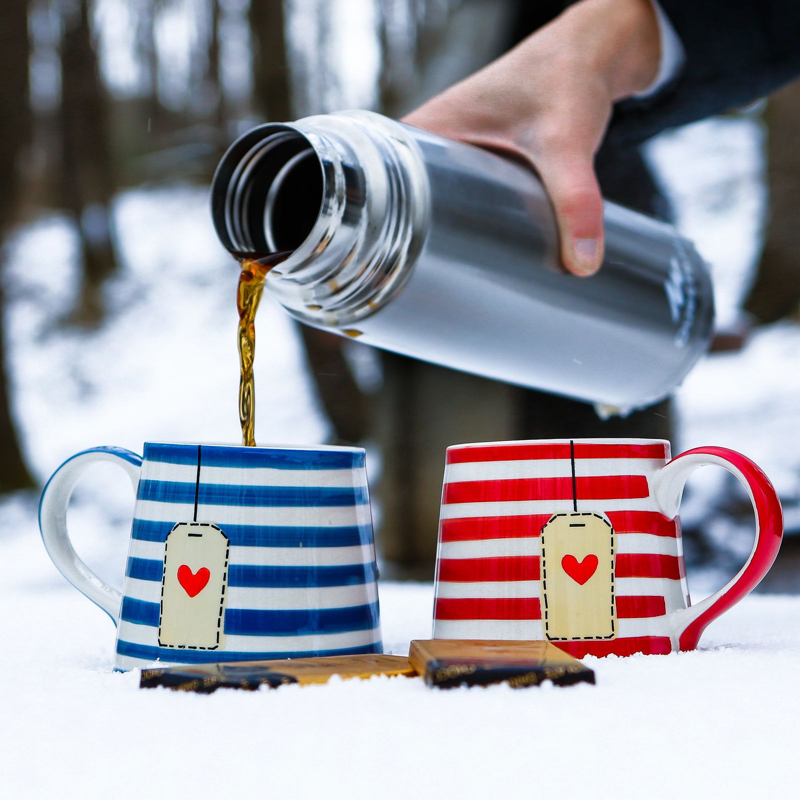 Hot drink poured from a flask into red and blue striped mugs standing in the snow