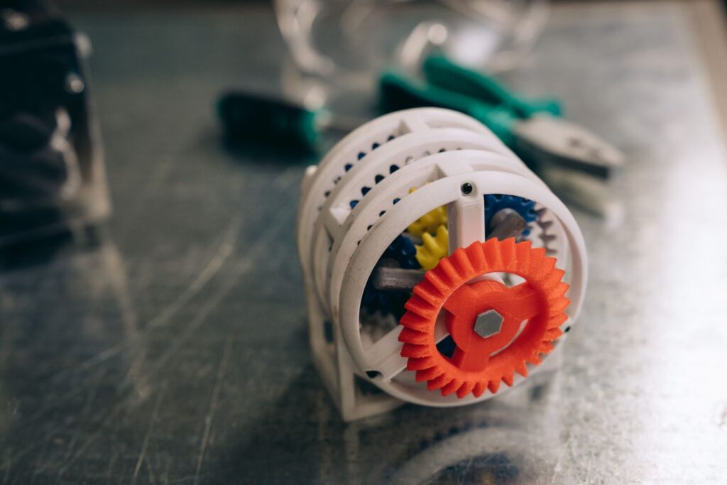 A 3D printed gear mechanism in white and orange on a metal bench