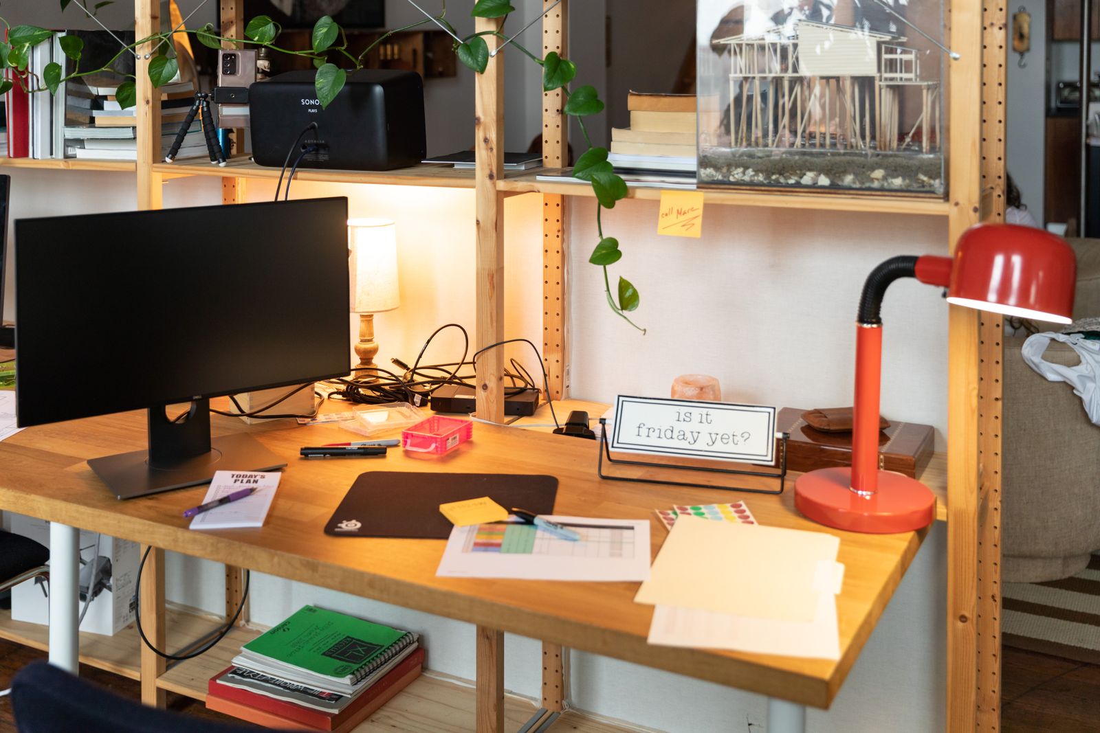 A red desk lamp on a wooden desk beside a monitor, notebooks and plants