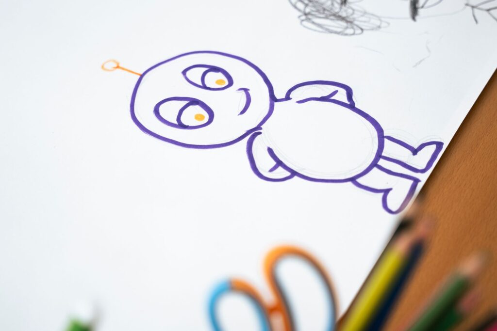 A cartoon character drawn in purple marker, with coloured pencils at the edge of the paper
