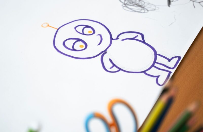 A cartoon character drawn in purple marker, with coloured pencils at the edge of the paper