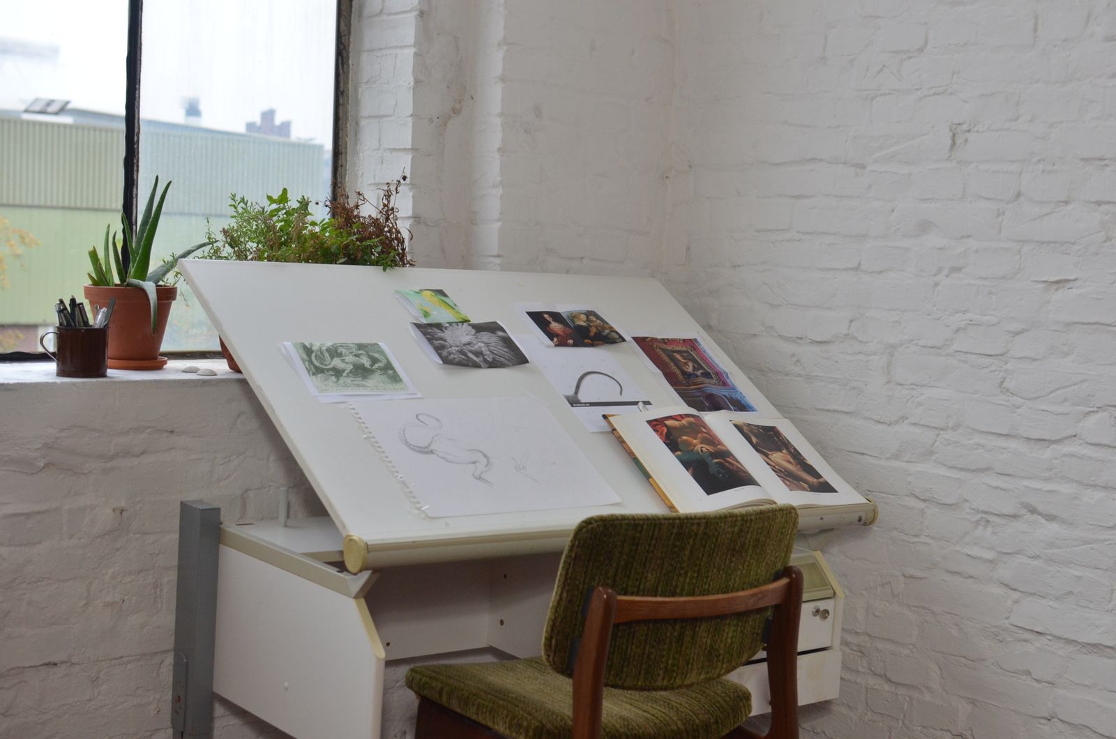 A small studio by a window: a tilted drawing table with pinned sketches, a chair and potted plants