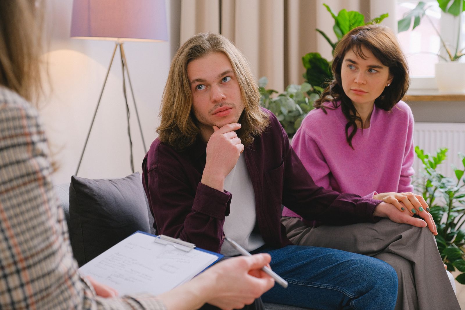 Two partners sitting side by side on a sofa, holding hands, while a therapist takes notes