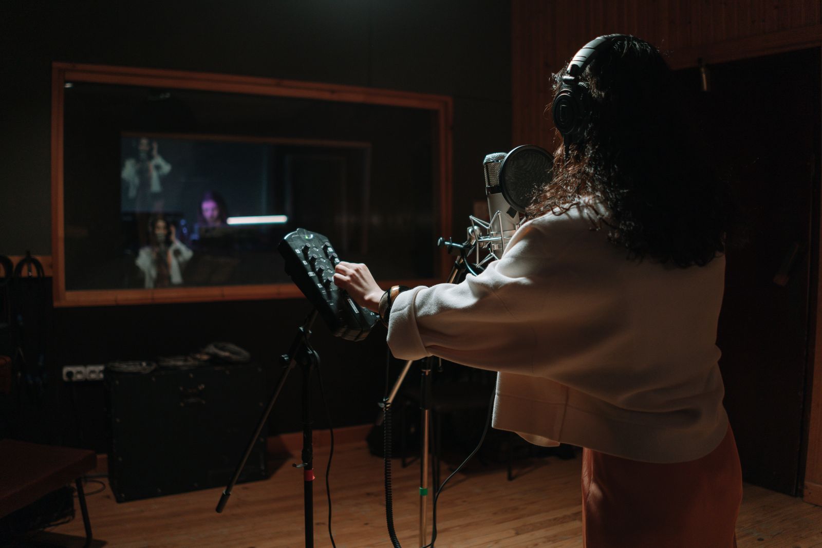 A singer in headphones seen from behind at a microphone, facing the control-room window
