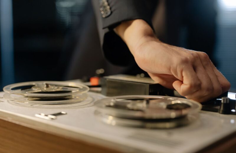 A hand pressing a key on an open-reel tape machine with two clear reels
