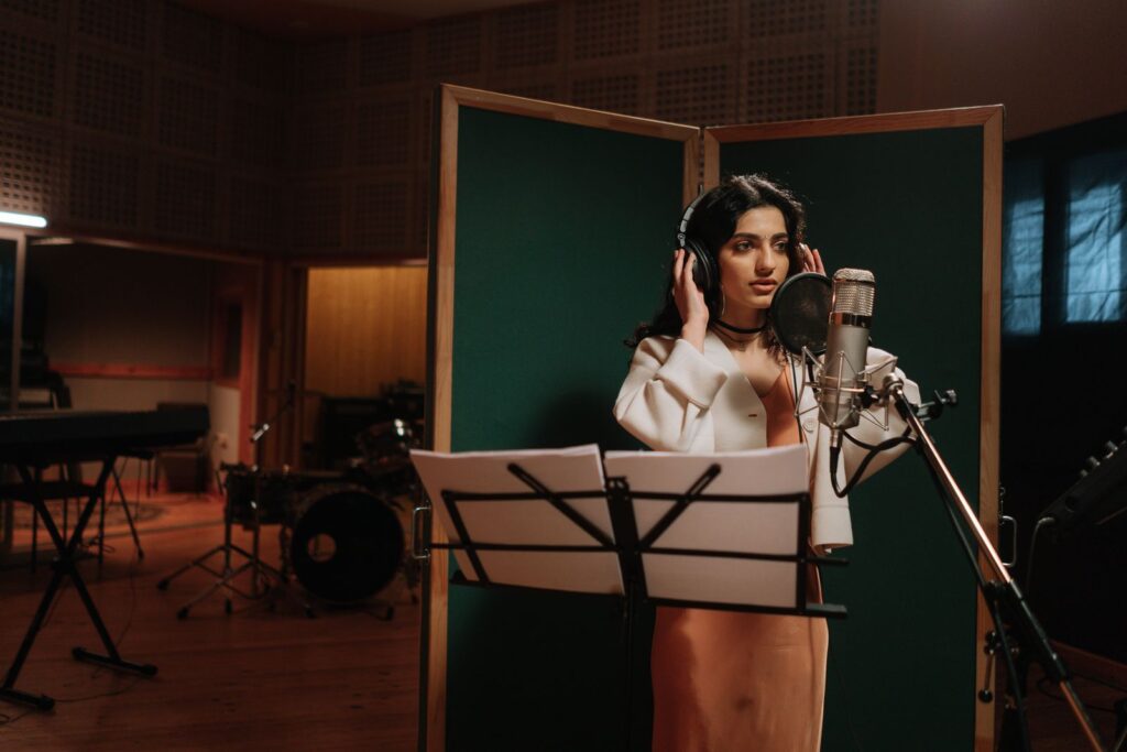 A singer in headphones at a microphone between two wooden screens in a live room, a drum kit behind her