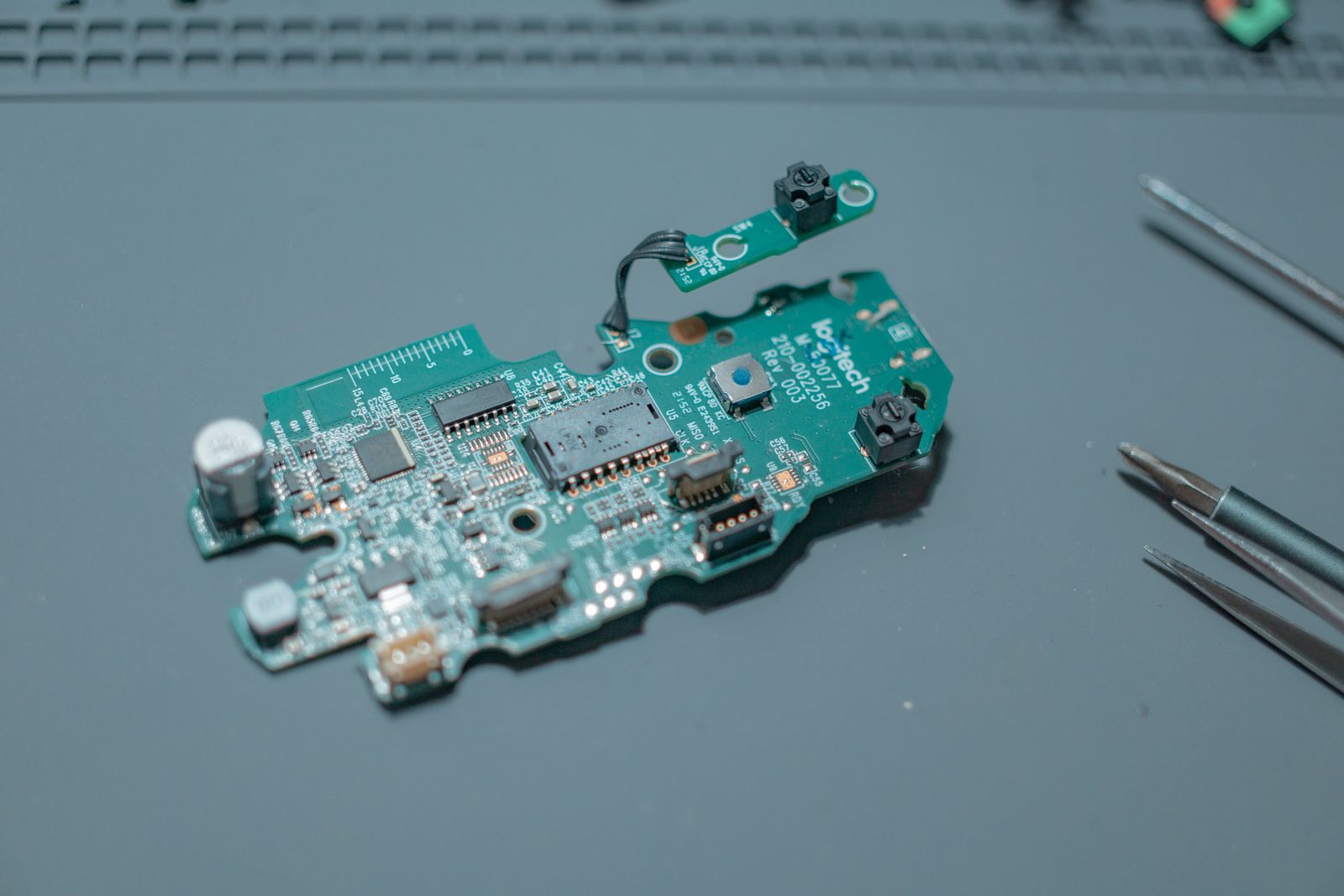 A green circuit board on a blue workbench with tweezers and small tools