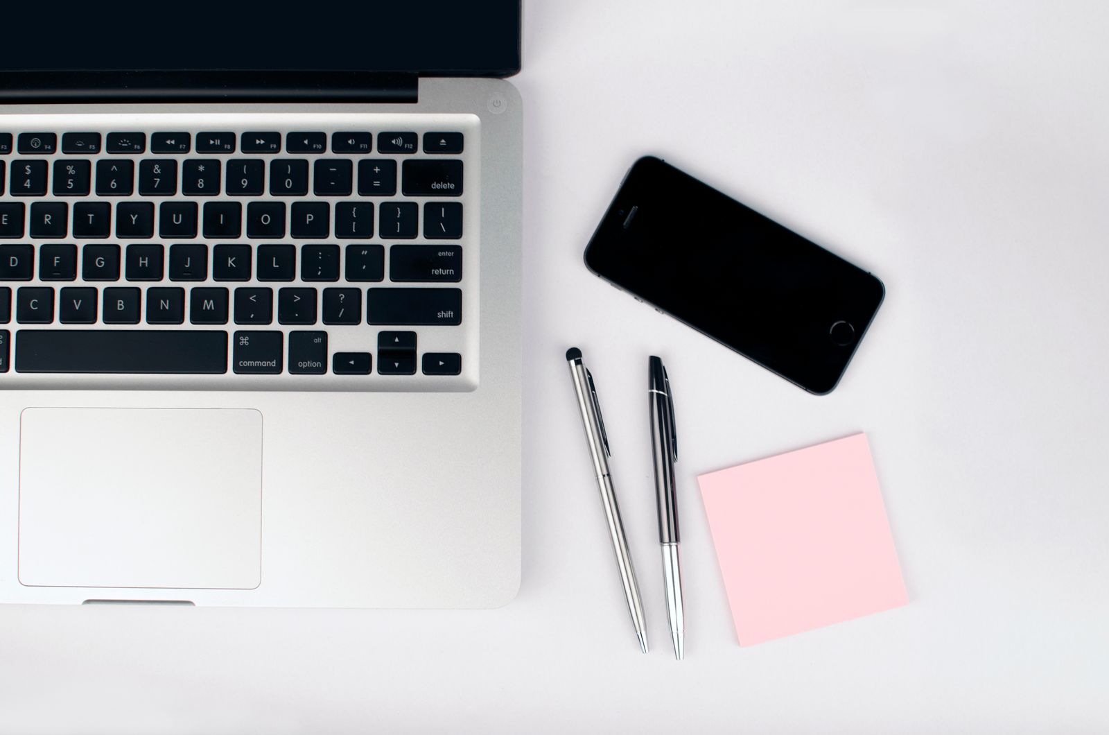An open silver laptop, a phone, two pens and a pink sticky note on a white desk, from above
