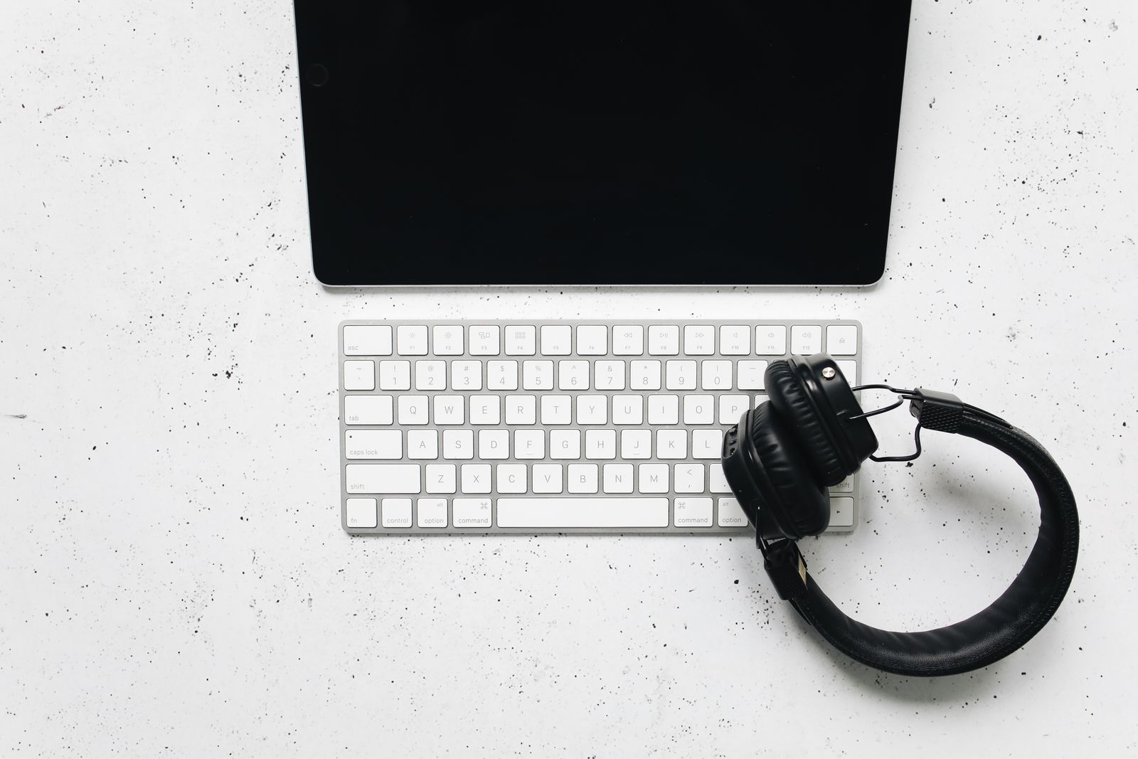 A tablet, a slim white keyboard and black over-ear headphones on a speckled white desk, seen from above