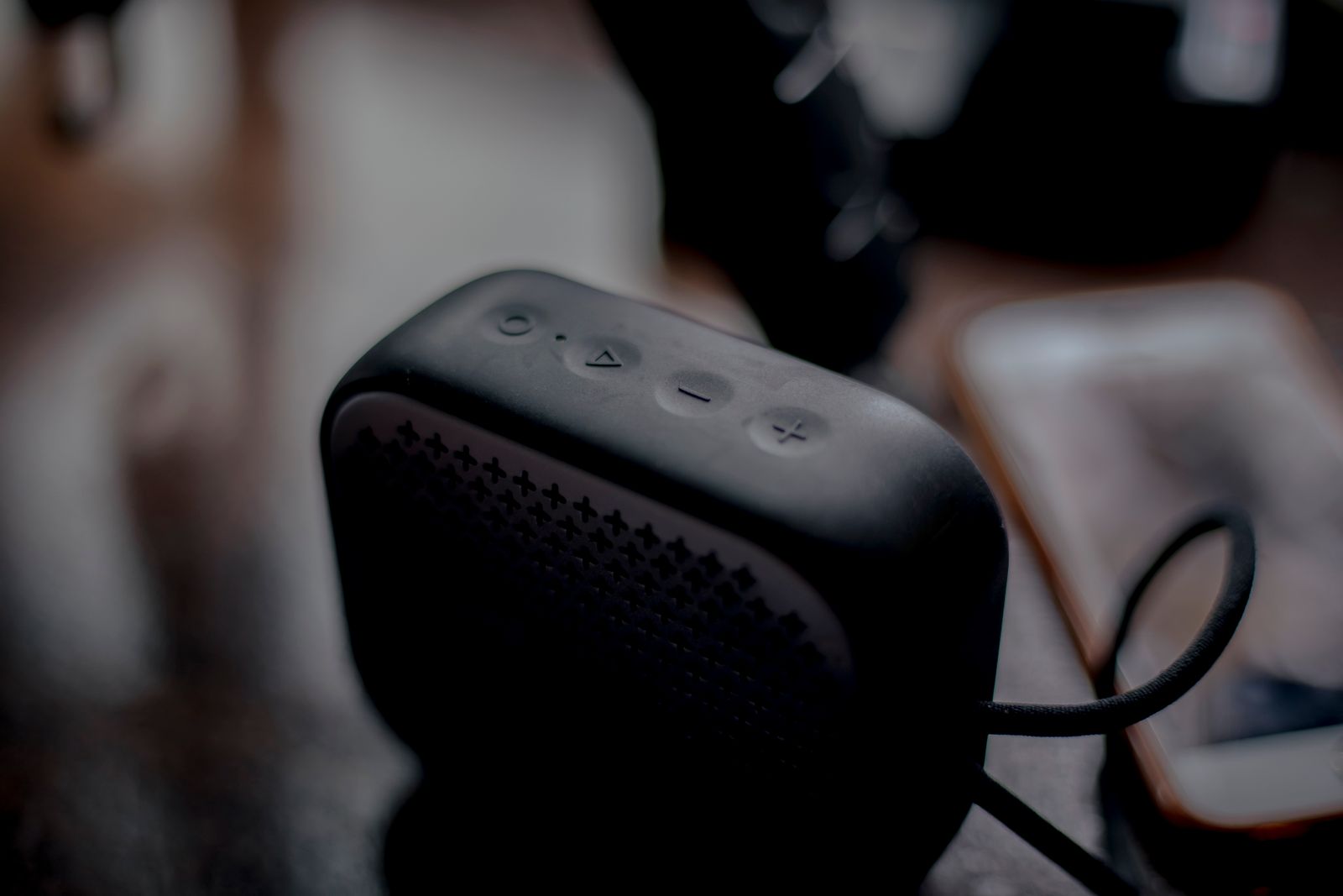 A small black portable speaker with round buttons on top, a phone blurred beside it