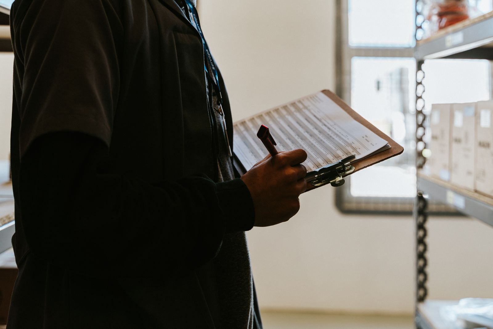 A person holding a clipboard and pen between warehouse shelves during a stock count