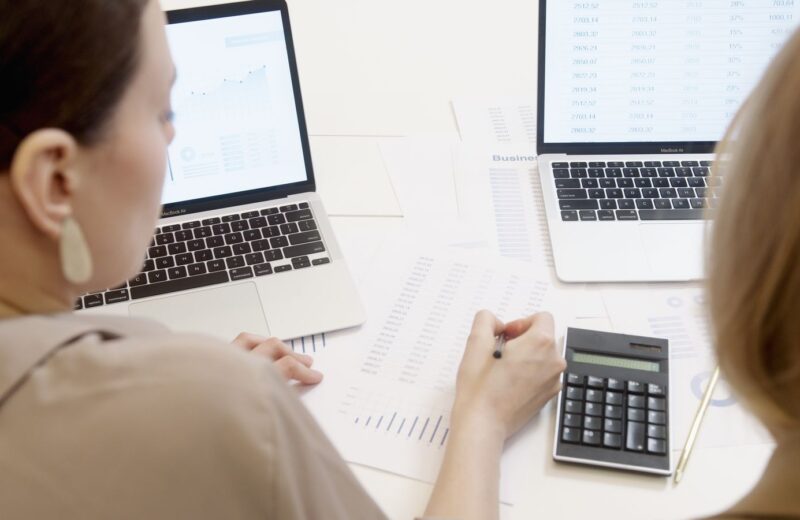 Two people at a desk with laptops, a calculator and printed figures