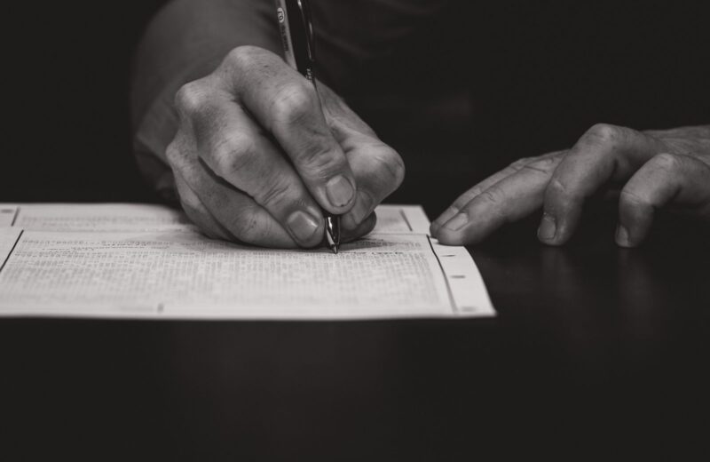 A hand signing a printed form with a pen, in black and white